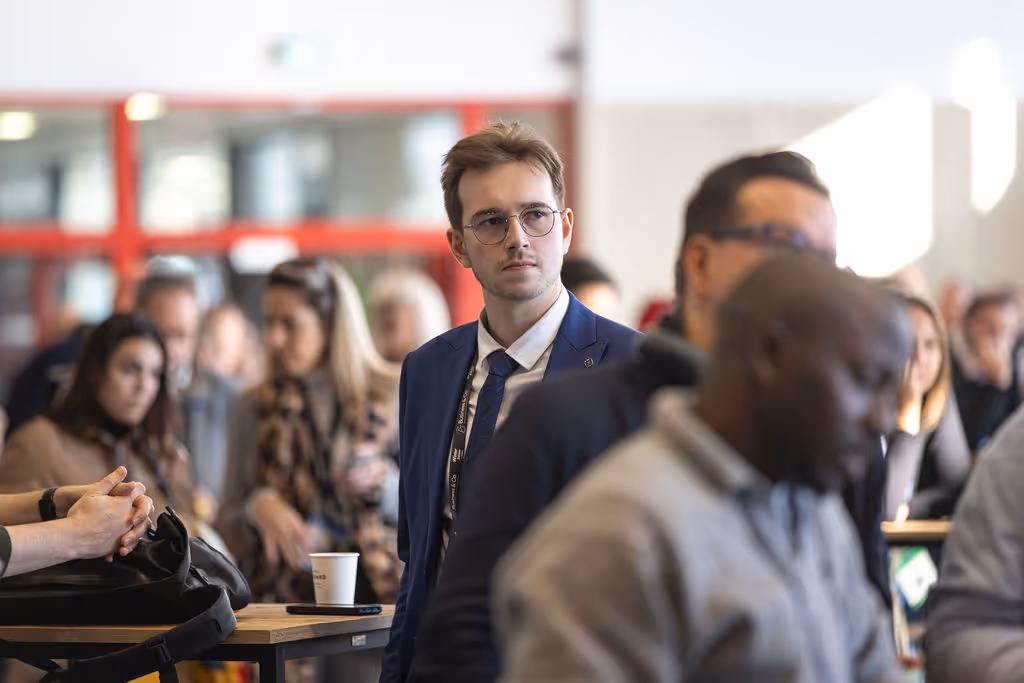A man in a blue suit and glasses stands attentively among a crowd indoors.