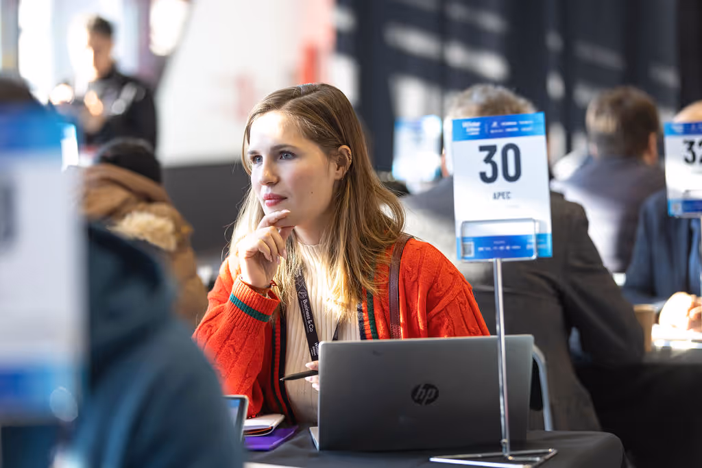 A woman in a red sweater sits at a table with a laptop, attending a conference.