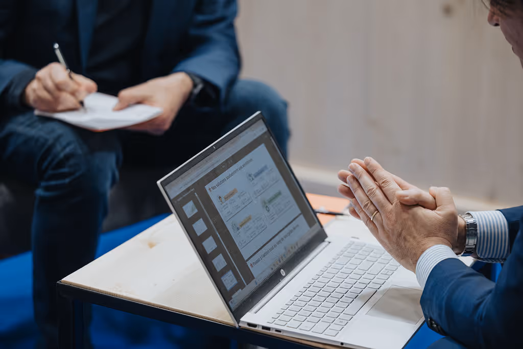 Two people in a meeting, one taking notes while the other gestures near a laptop.