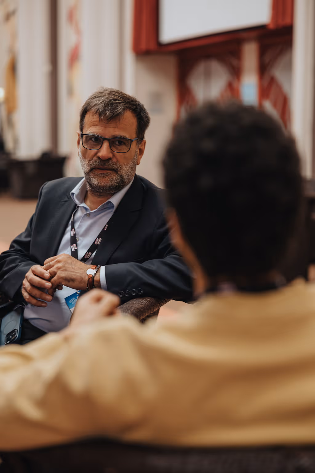 Two people sitting and talking in a room with red curtains.