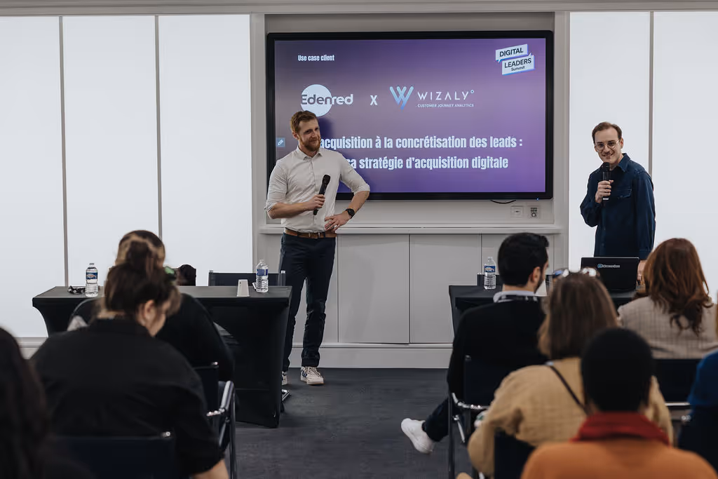 Two men stand and speak at the front of a room with a presentation screen behind them.