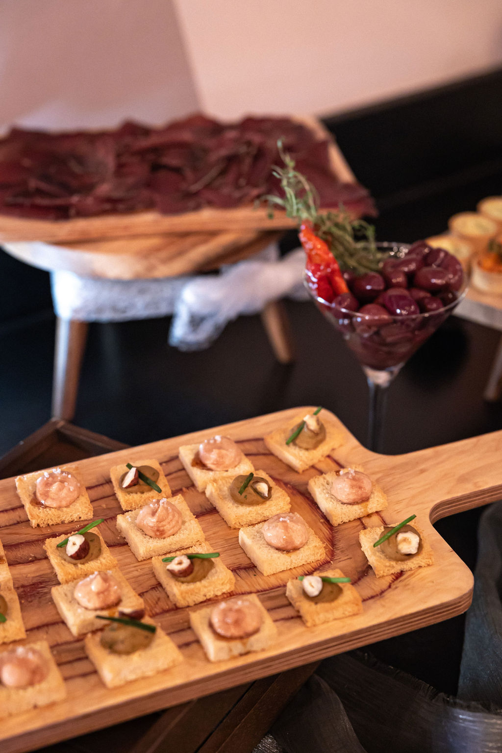 Assorted appetizers on a wooden tray with cheese and herbs, accompanied by a glass of olives.