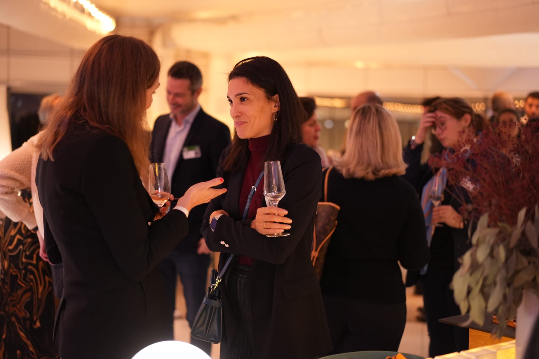 Two women in conversation, holding wine glasses at a social gathering. Other people in the background.