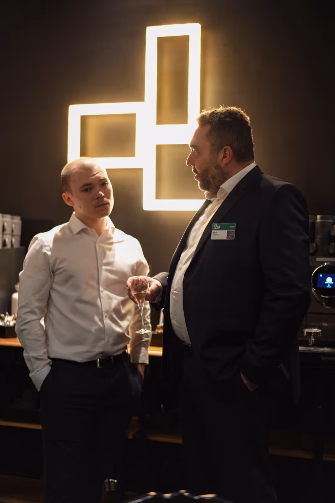 Two men in formal attire talking indoors, one holding a wine glass, standing under a lit sign.