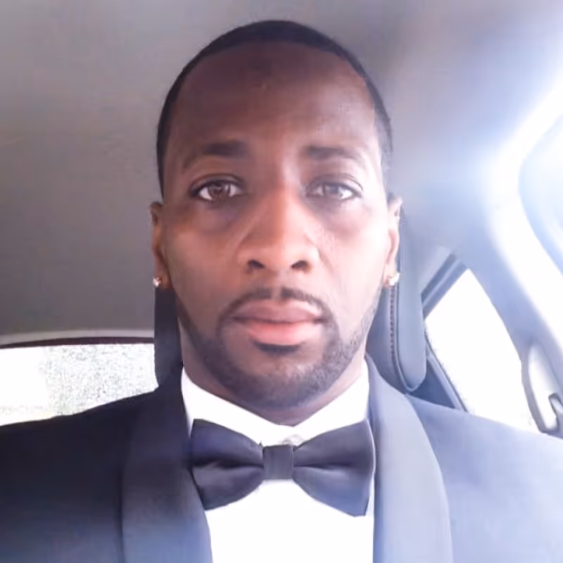 Man in a tuxedo sitting inside a car looking directly at the camera.