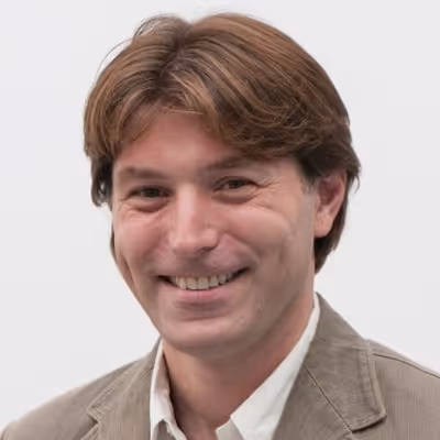 A person with brown hair smiles while wearing a beige jacket over a white shirt.