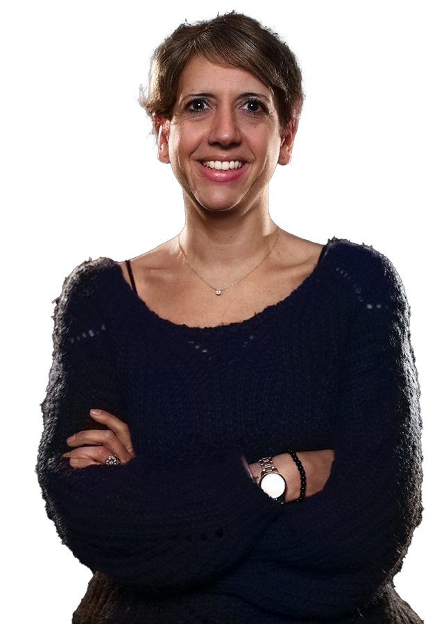 Smiling person with short hair and crossed arms, wearing a dark sweater, stands facing forward.