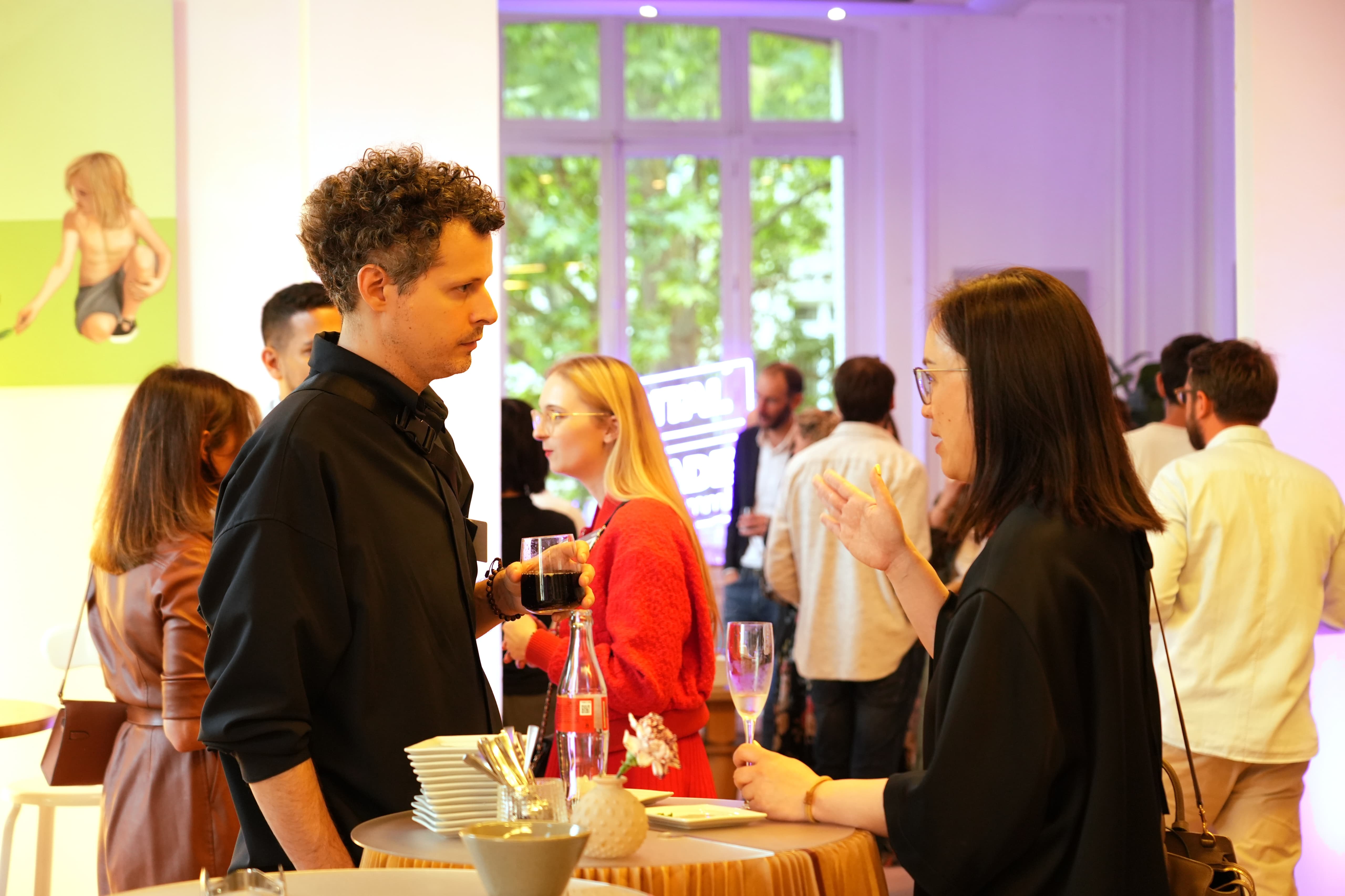 A man and woman converse at a reception, each holding a drink, with others in the background.