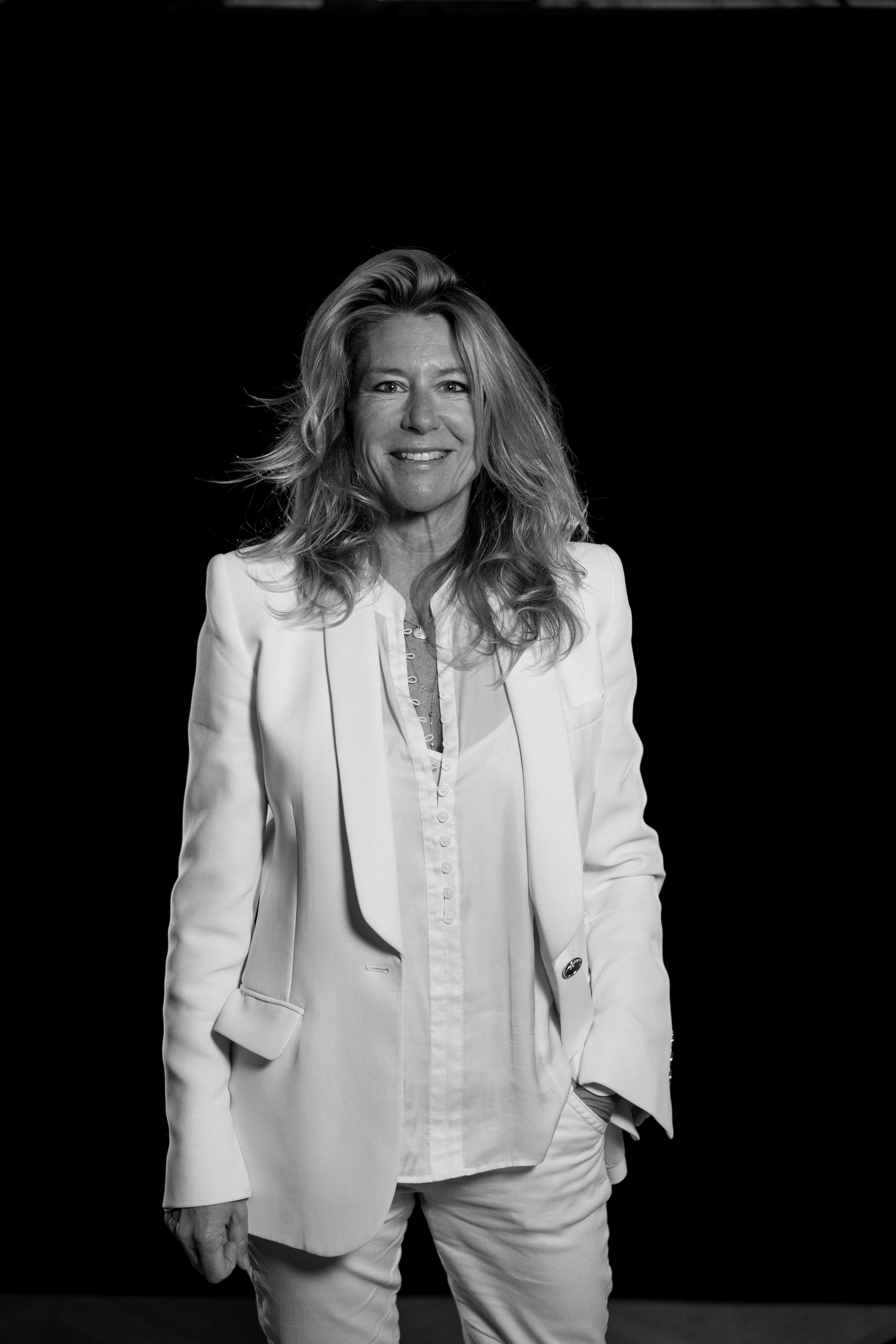 A woman in a white suit stands and smiles against a black background.