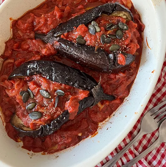 Aubergines rôties à l’ottomane garnies de sauce tomate maison, servies dans un plat blanc avec graines de courge, sur une nappe à rayures rouges