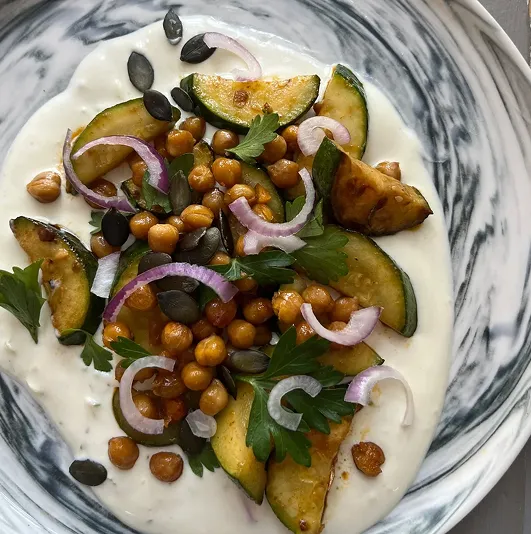 Courgettes grillées et pois chiches rôtis sur lit de sauce feta citronnée, garnis d’oignons rouges, persil et graines, servis dans une assiette marbrée