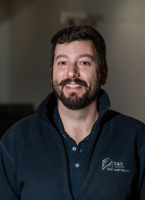 Smiling man with glasses wearing a dark blue D&S Smart Housing jacket indoors.