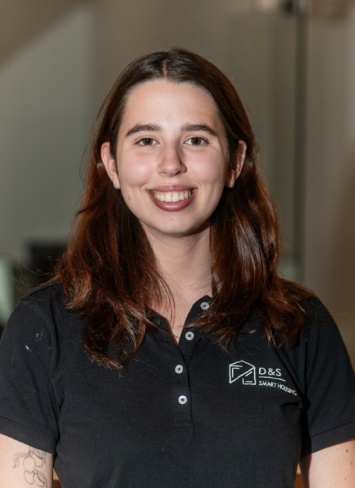 Smiling young woman with long brown hair wearing a black cropped polo shirt and blue jeans standing indoors.