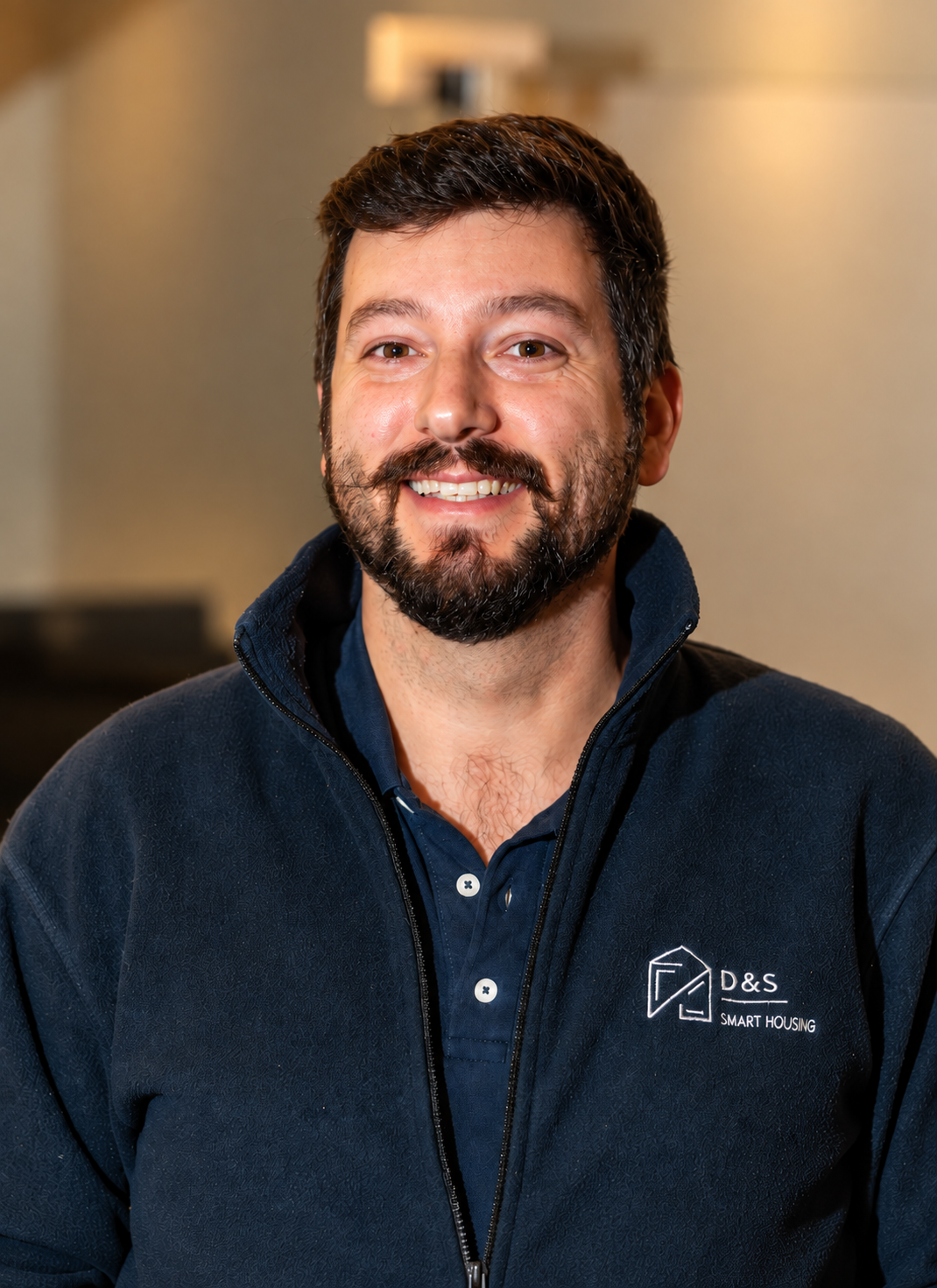 Smiling man with glasses wearing a dark blue D&S Smart Housing jacket indoors.