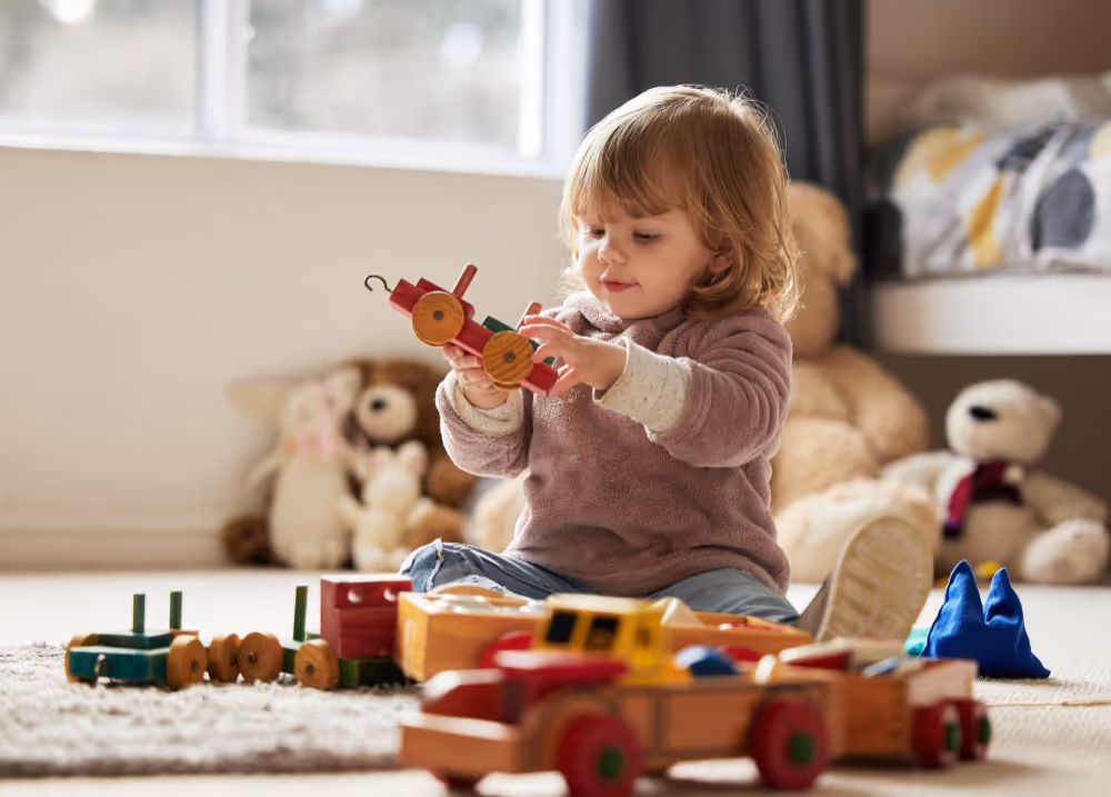 Kleines Kind, das in einem hellen Zimmer auf dem Boden sitzt und mit einem hölzernen Spielzeugzug spielt.