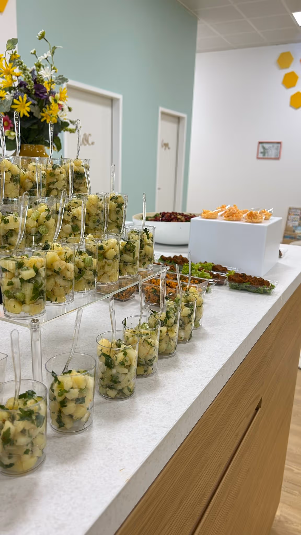 Gläser mit Kartoffelsalat und Kräutern auf Buffet mit weiteren Snacks auf Tischen.