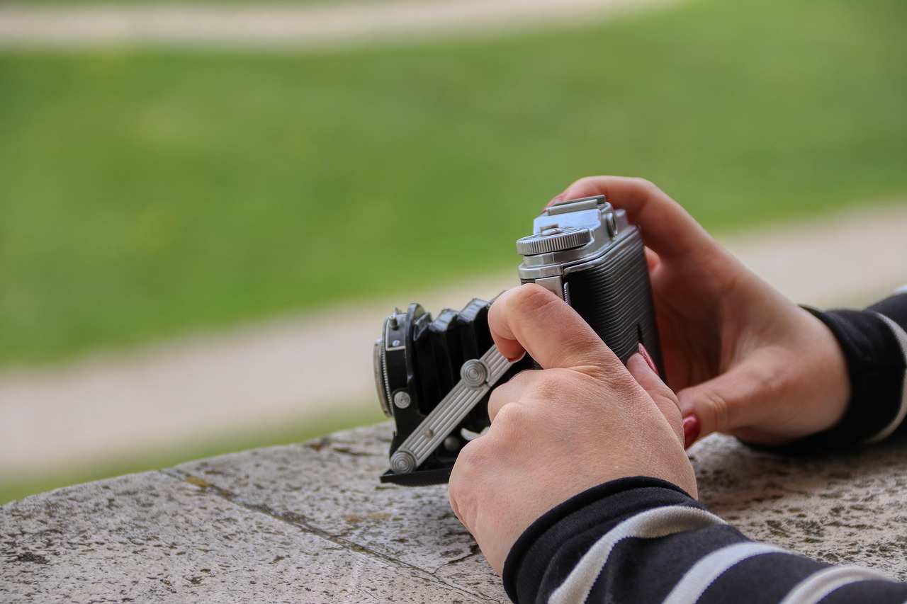 Hands holding a vintage folding camera resting on a stone ledge, with a softly blurred green outdoor background