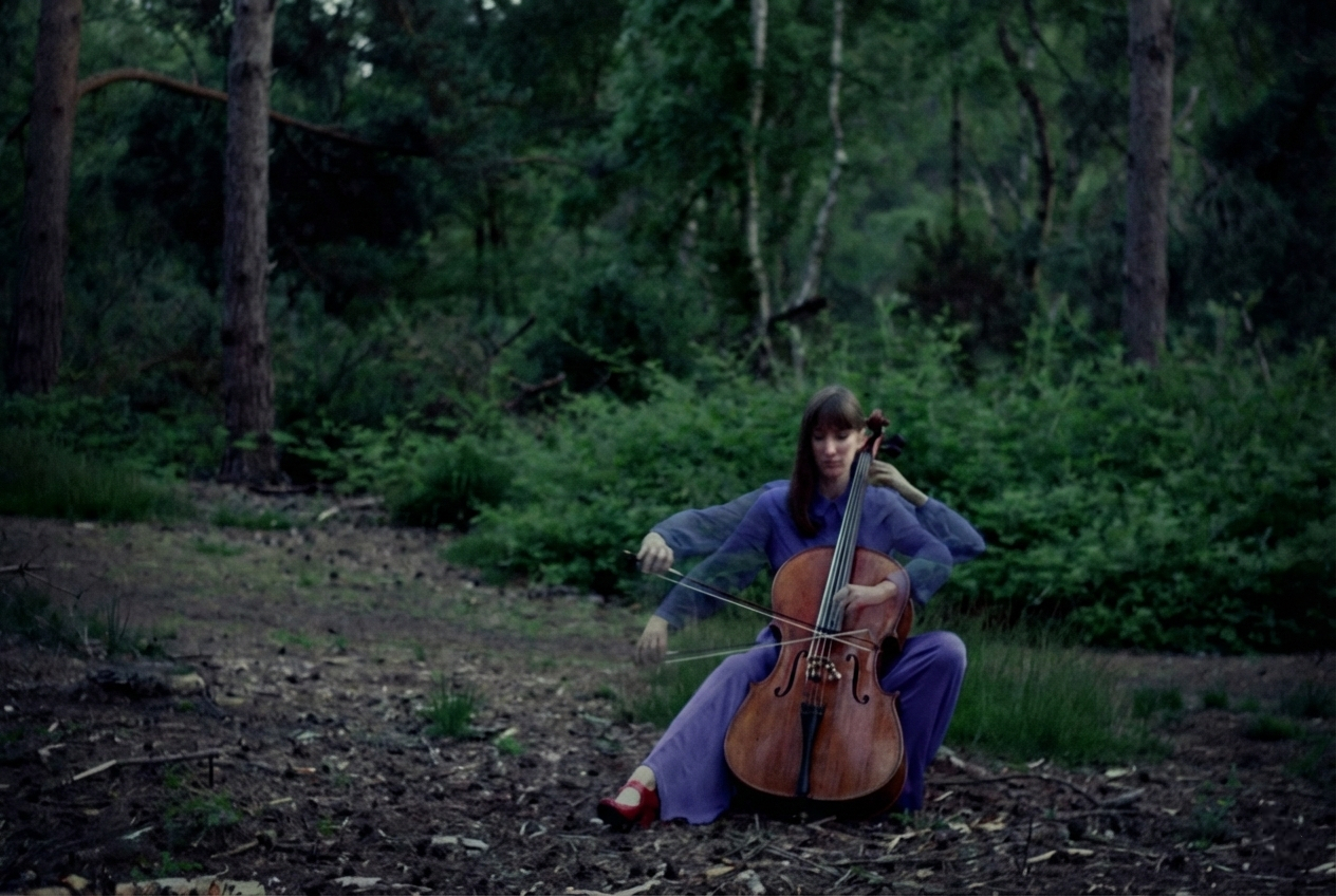 Naomi in a purple outfit sitting on the forest floor playing a cello, with slight motion blur in her arms against a backdrop of dense green trees