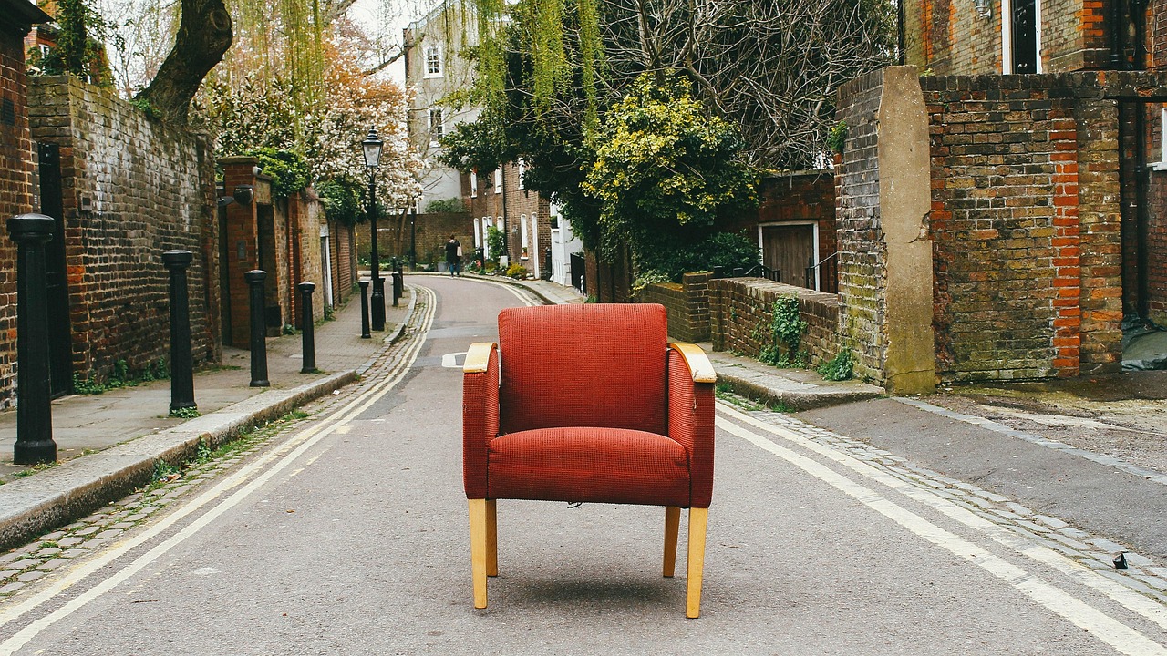 A single red armchair placed in the middle of a quiet residential street, surrounded by brick walls and trees
