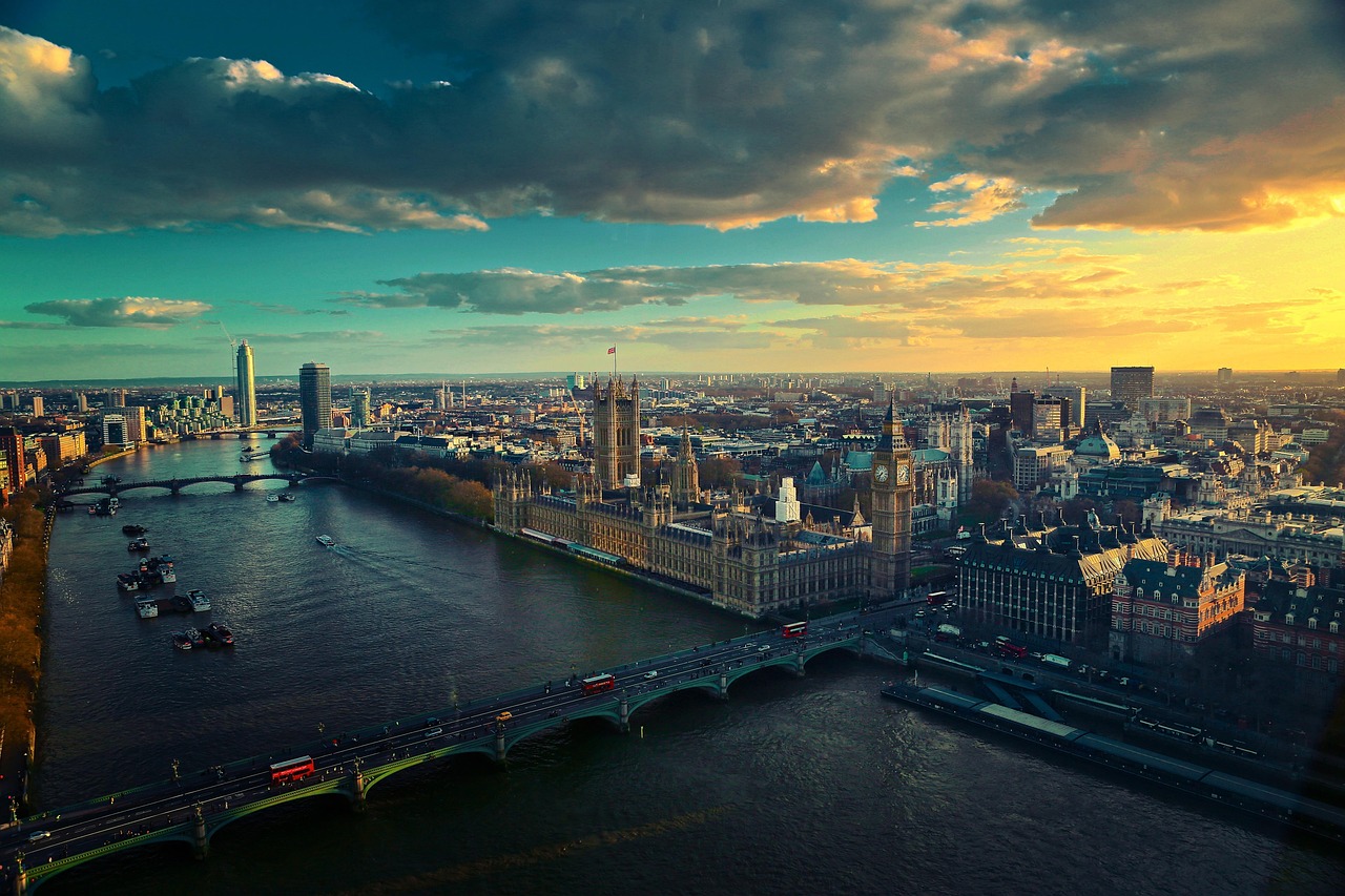 A wide aerial view of London at dusk, showing the River Thames, the Houses of Parliament, Big Ben, and surrounding city skyline under a dramatic evening sky