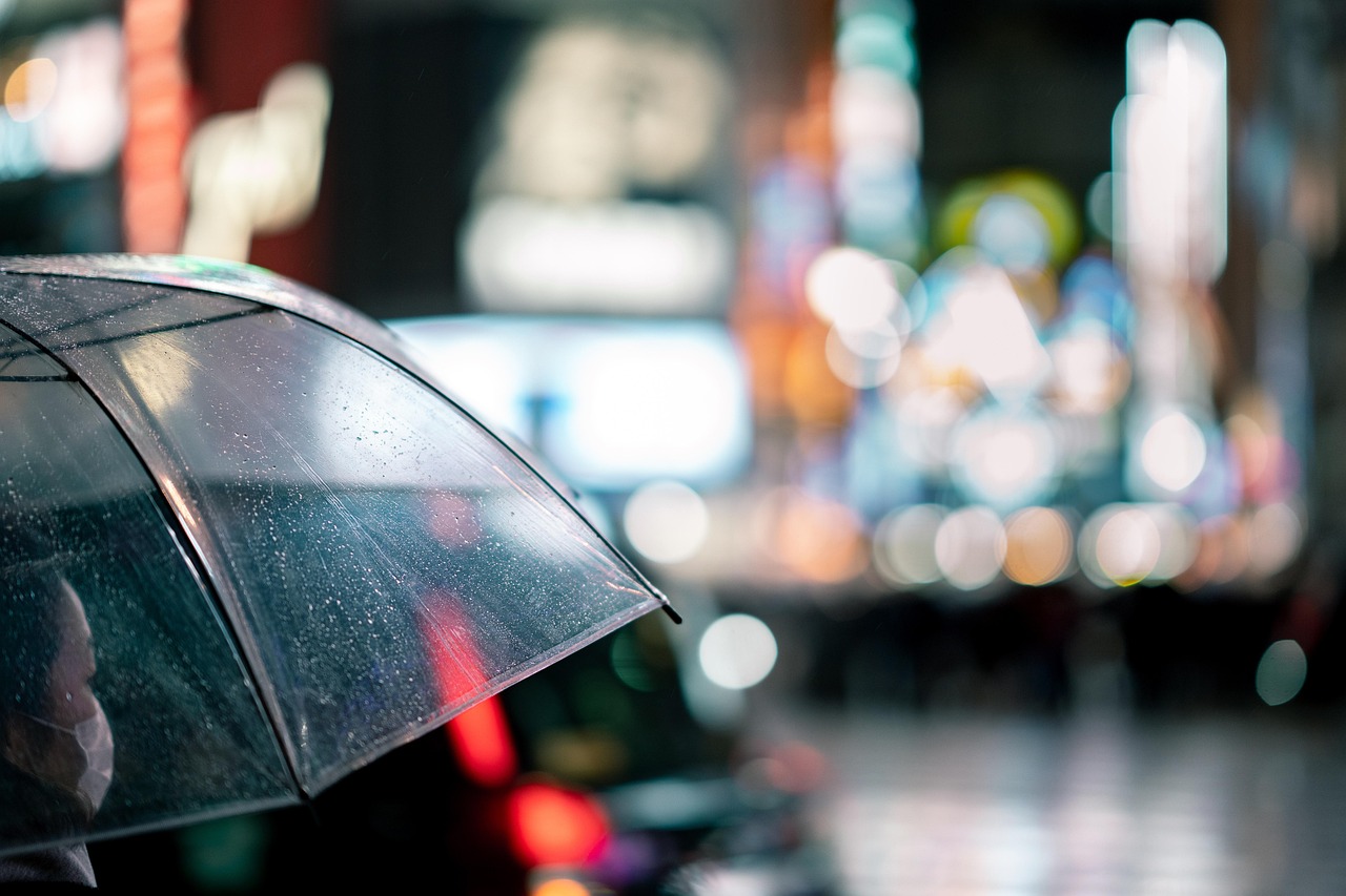 A person sheltering beneath a clear umbrella on a rainy city street, with blurred lights in the background.