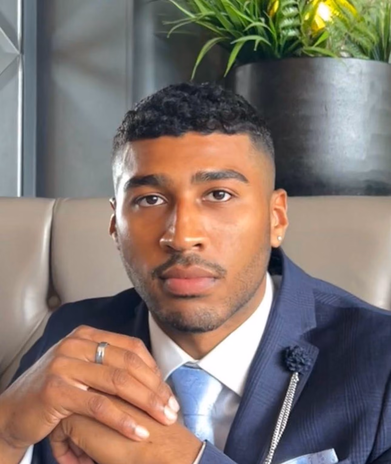 Man in a blue suit with light blue tie and silver pin, sitting with hands clasped, looking forward.
