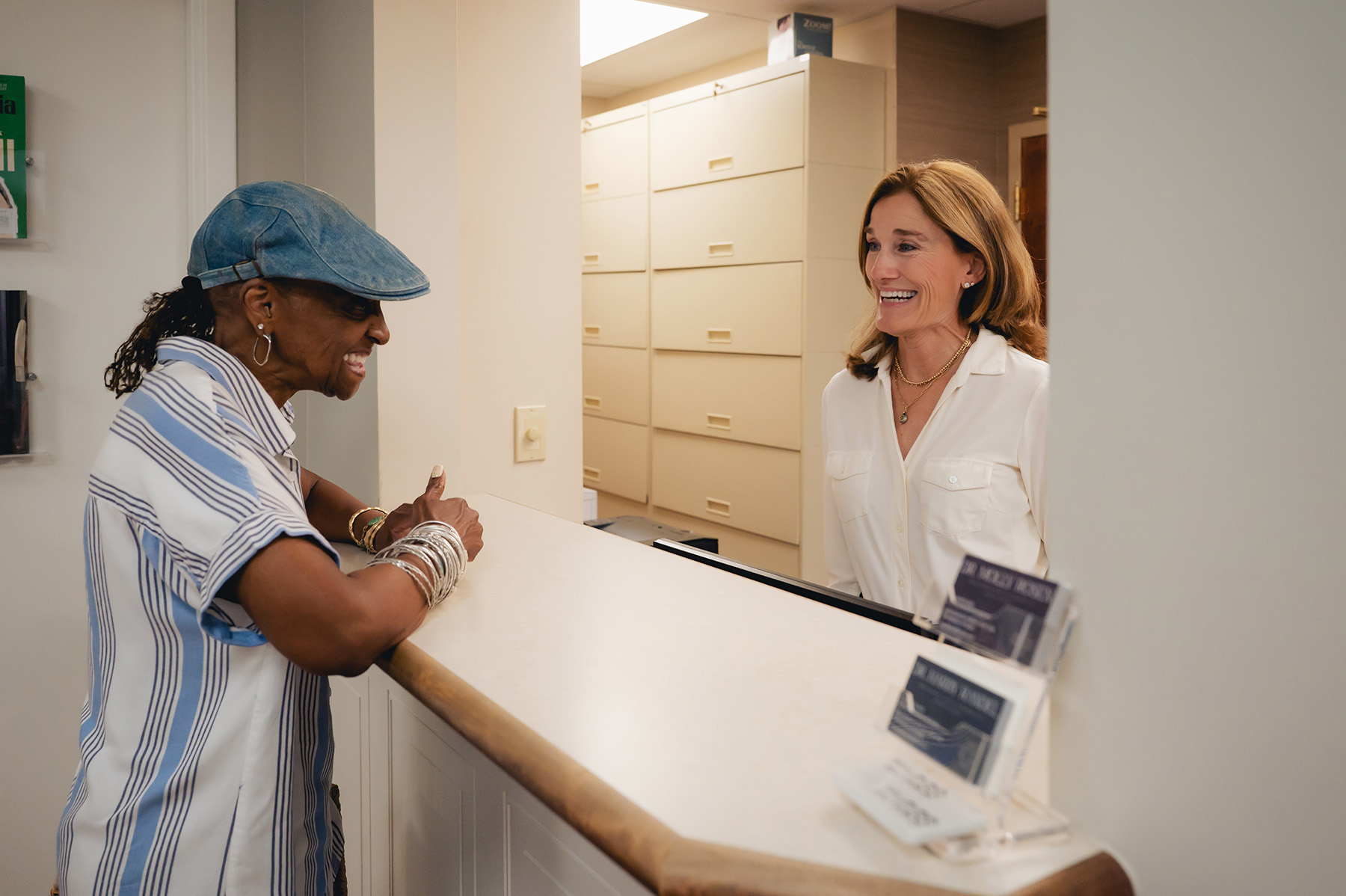 A woman speaks and smiles with a receptionist across a counter in a well-lit dentist’s office, with filing cabinets in the background.