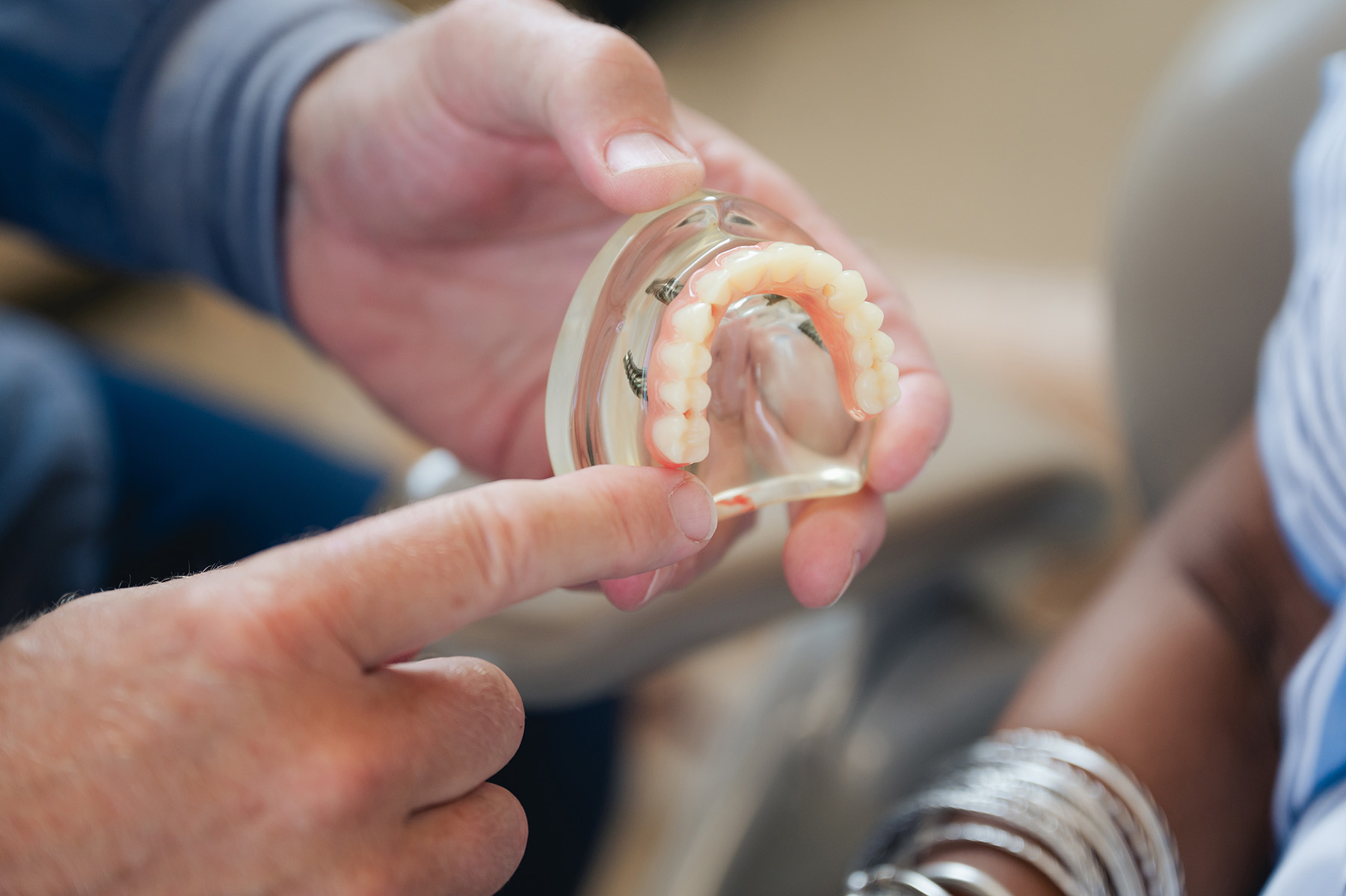 A dentist holds and points to a dental implant model showing artificial teeth and gums, explaining dental treatment to another individual.