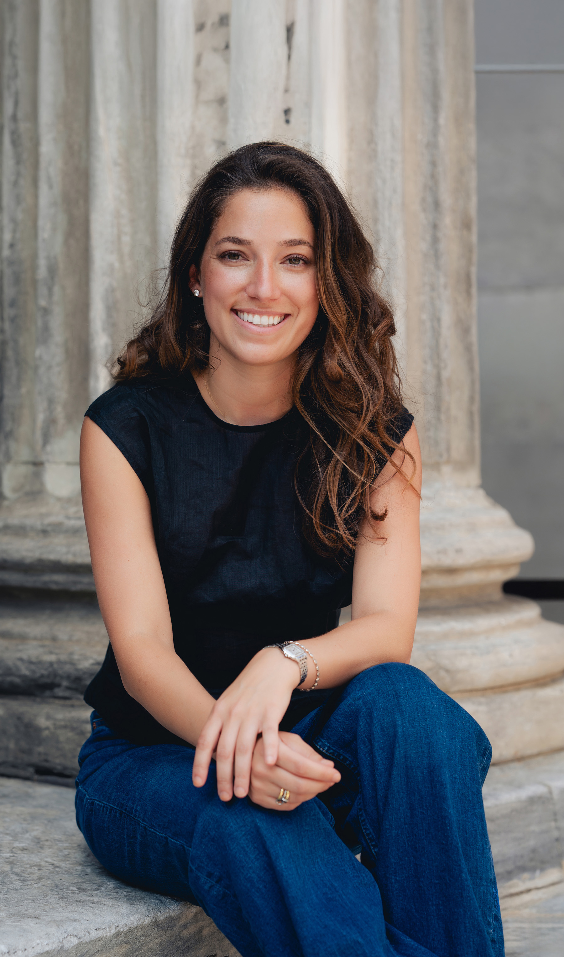 A woman with long brown hair, wearing a black top and blue jeans, sits on stone steps and smiles at the camera, showcasing her bright dentist-approved smile. Stone columns are visible in the background.