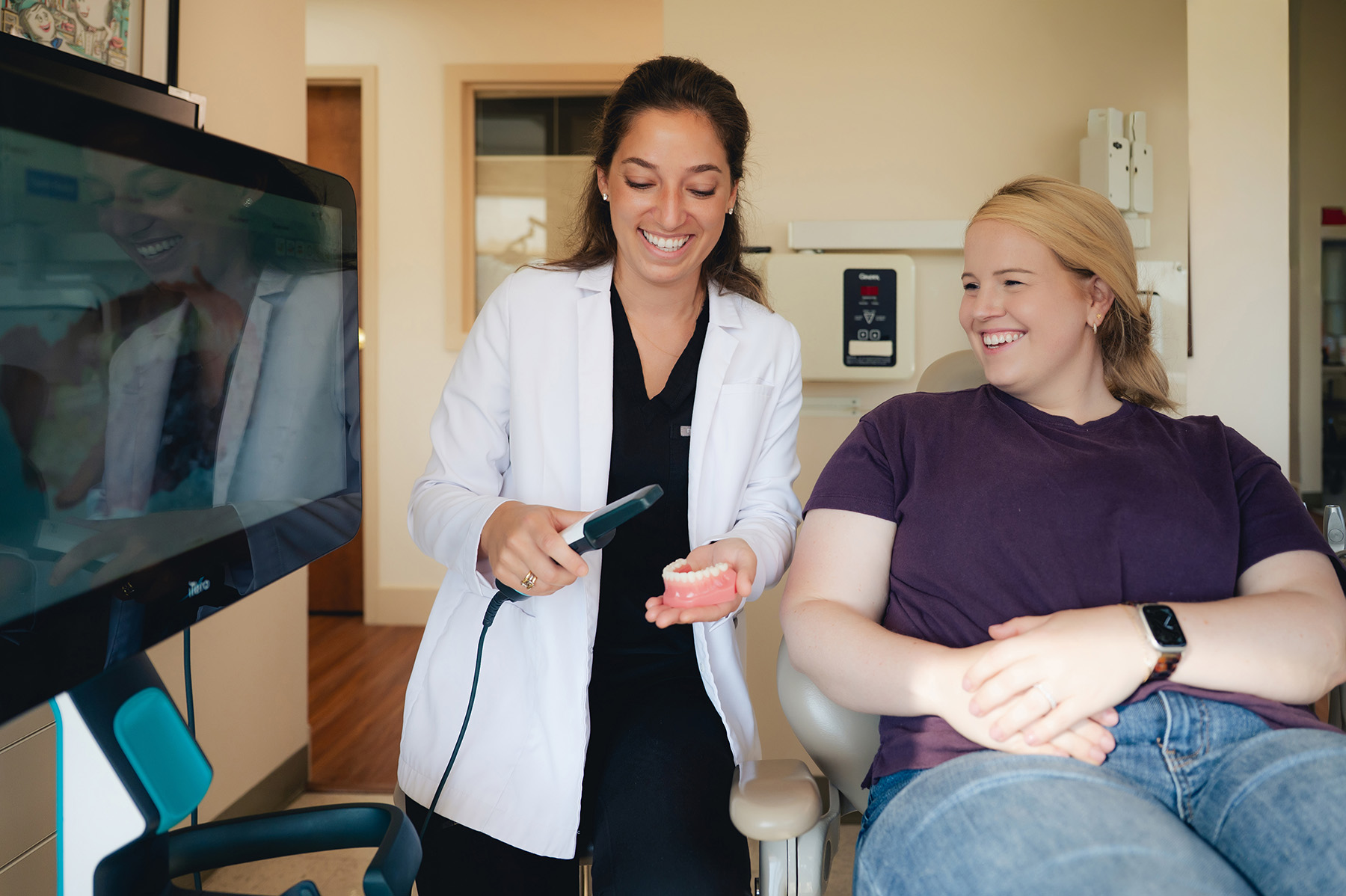 Female dentist demonstrating dental tool on a model of teeth to a smiling patient seated in a dental chair.