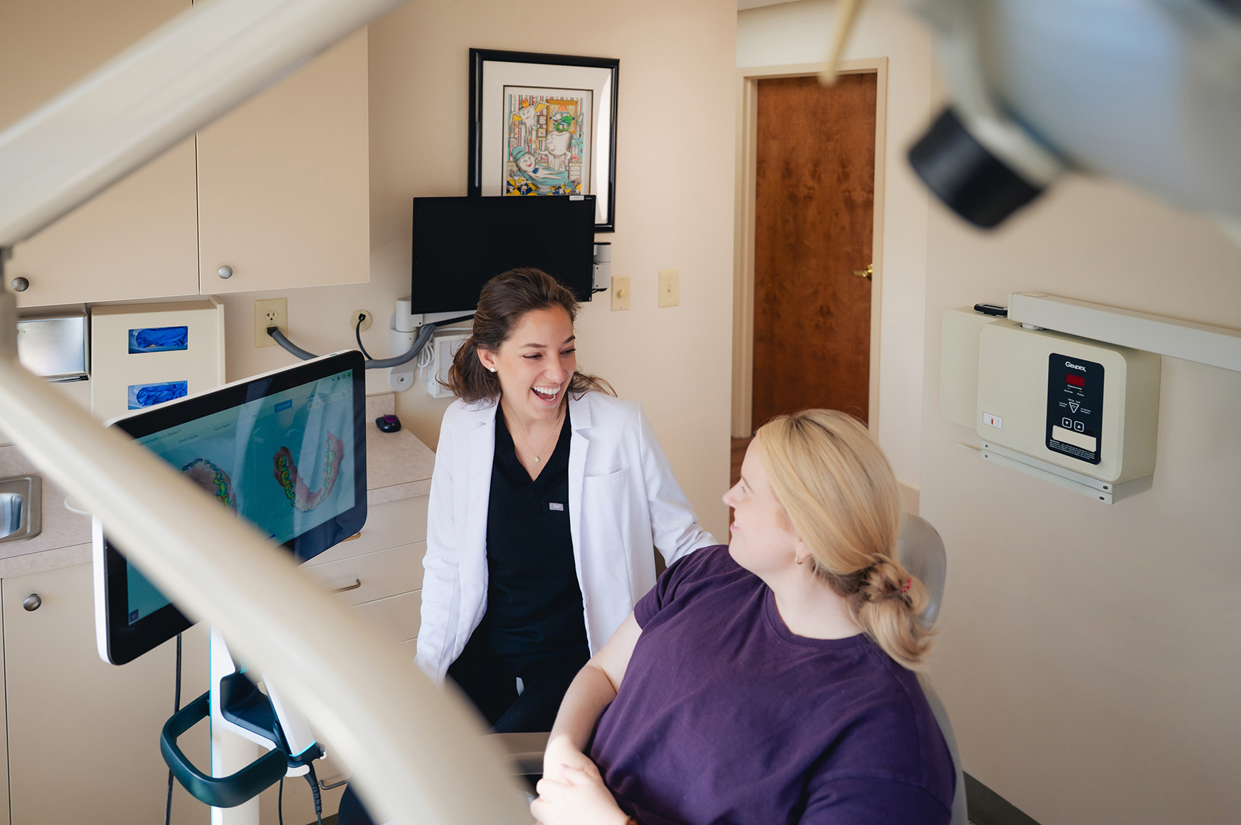 A dentist in a white coat talks with a patient seated in a dental chair in a modern dentist examination room.