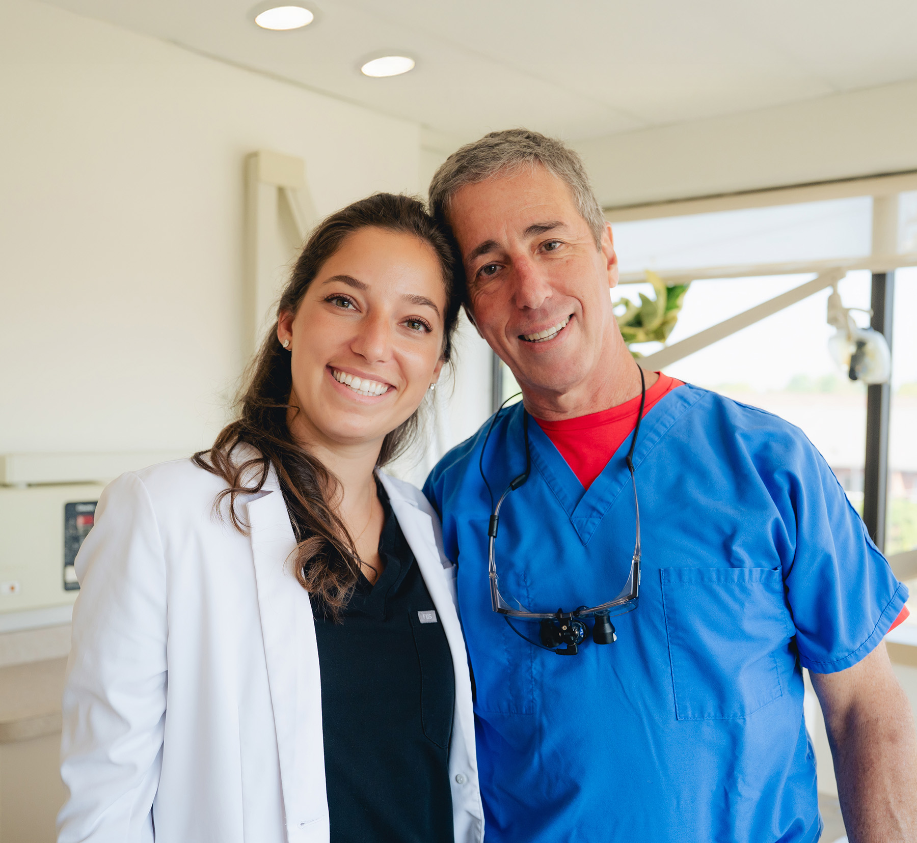 Two healthcare professionals, a dentist in blue scrubs with dental loupes and another in a white coat, stand side by side smiling in a bright clinic room.