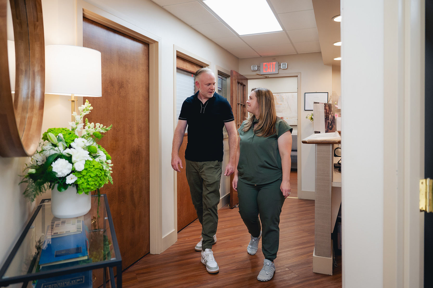A man and a woman walk and talk together in the hallway of a dentist’s office with wooden floors and doors, under bright lighting.