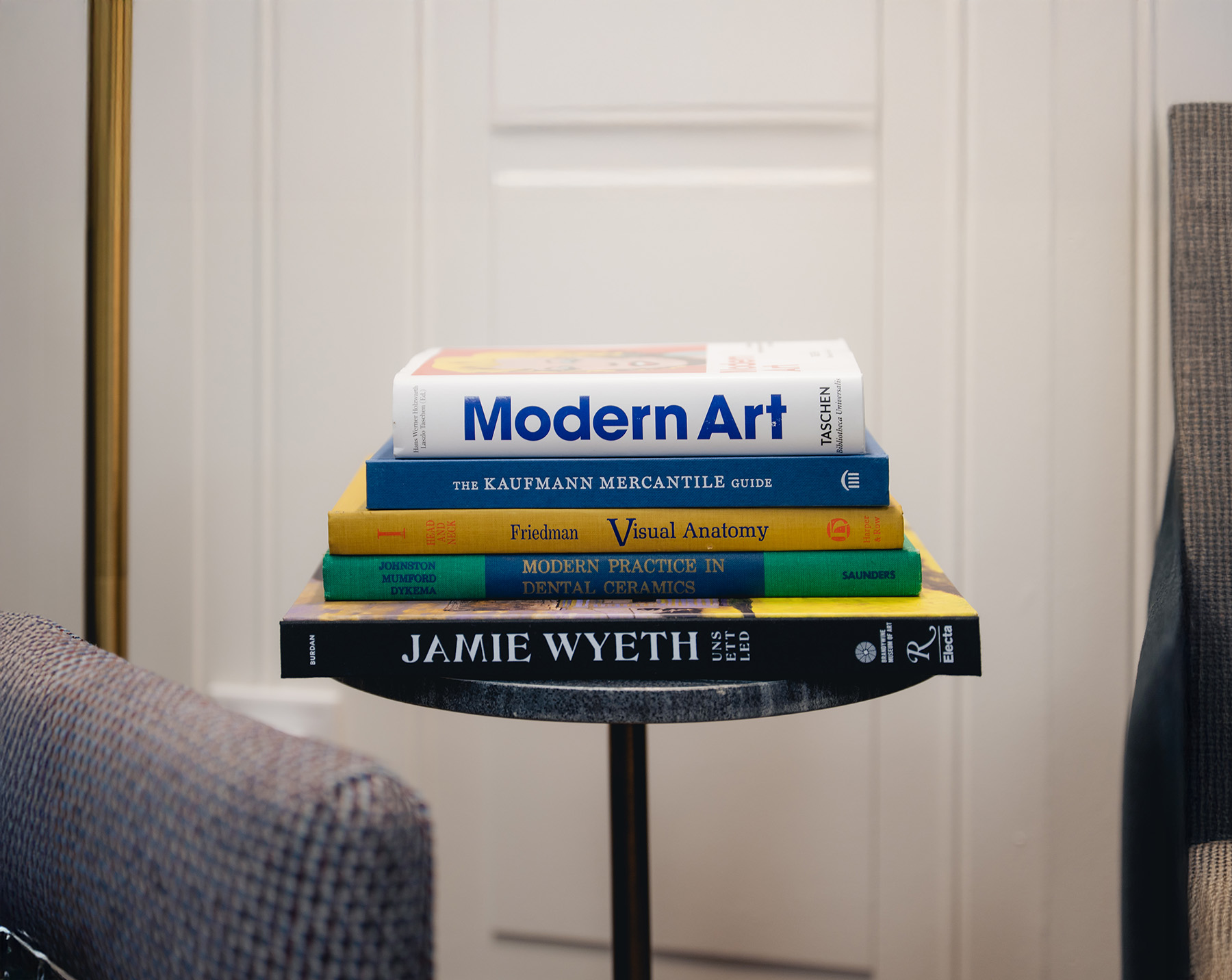 A stack of five books on a small table, including Modern Art, The Kaufmann Mercantile Guide, Visual Anatomy, Modern Practices in Dental Ceramics—a must-read for any dentist—and Jamie Wyeth.