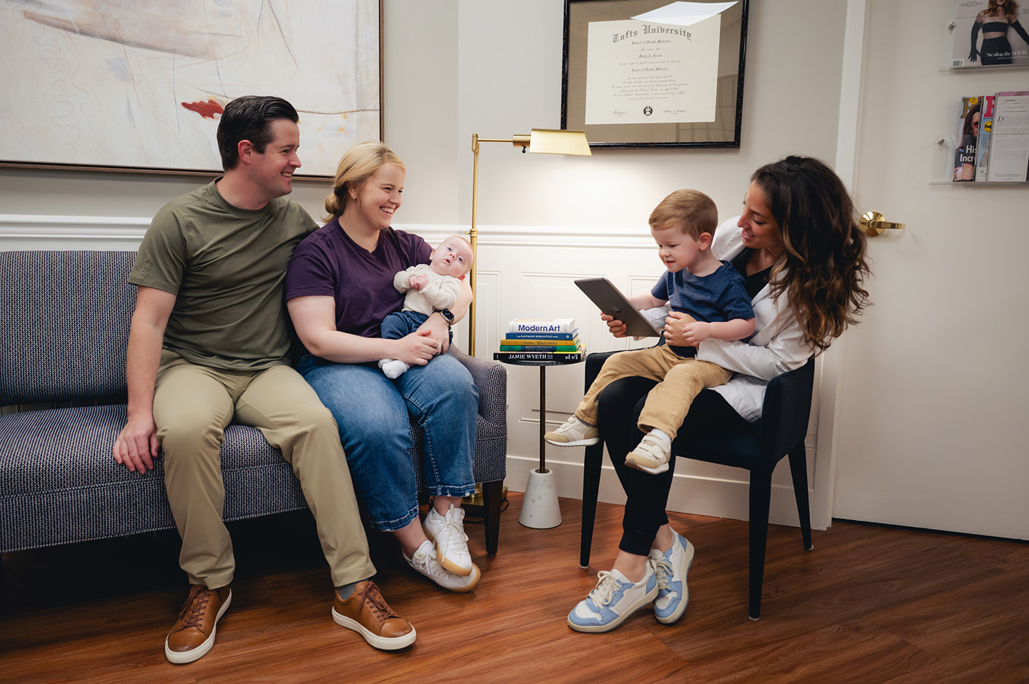 Happy family with baby sitting on a couch and a woman with a child on her lap using a tablet in a cozy room.