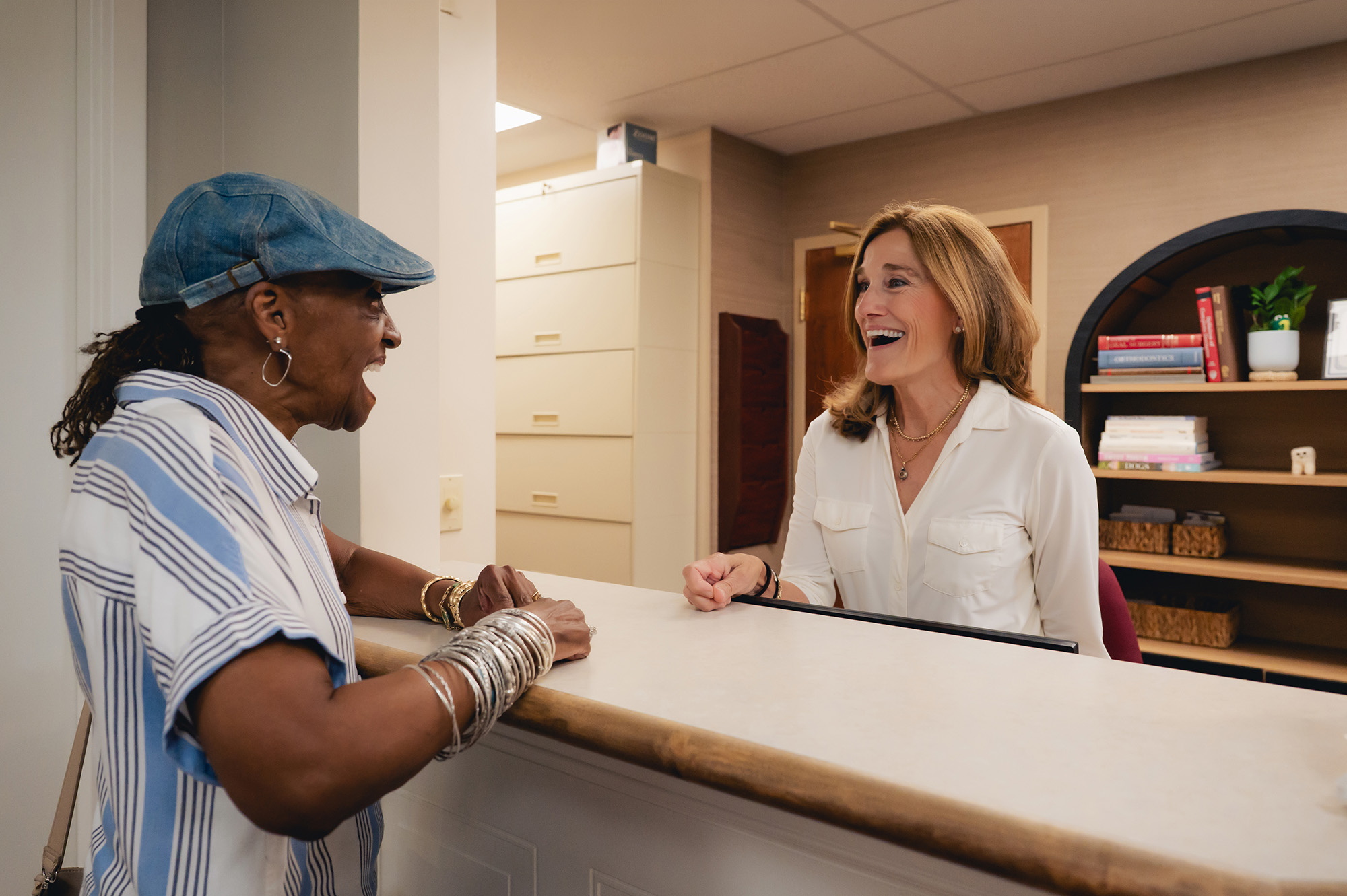 Two women smiling and talking across a reception desk in a well-lit office.