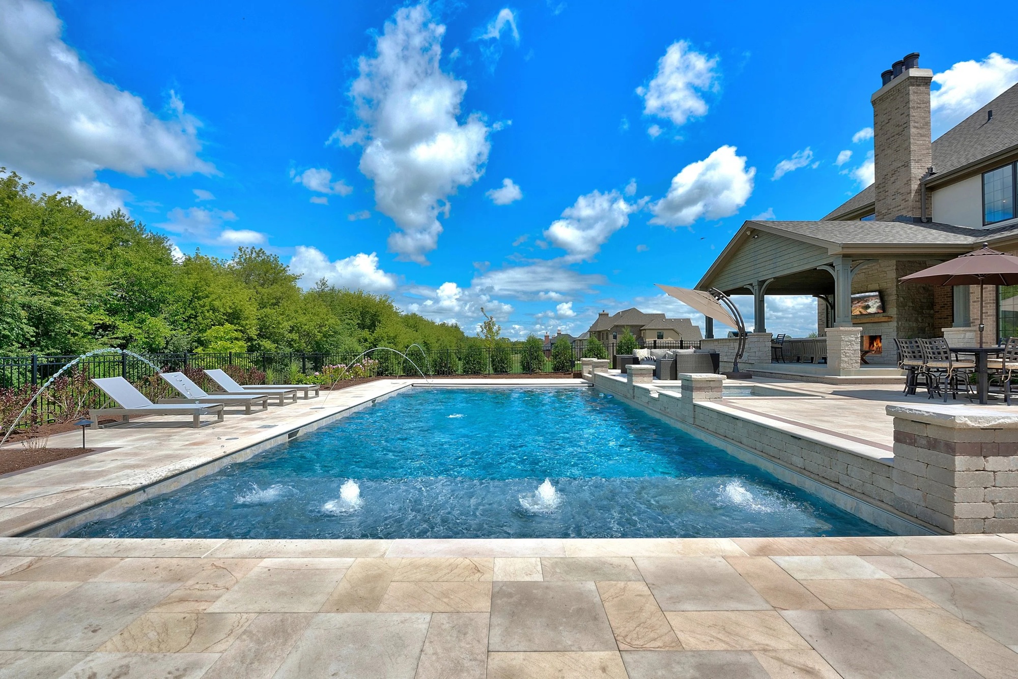 A large, luxurious rectangular custom-built swimming pool with integrated water features and a spacious stone patio, set against a blue sky. This exemplifies Mooresville Hire Custom Pool Construction by Craft Master Pools and Spas.