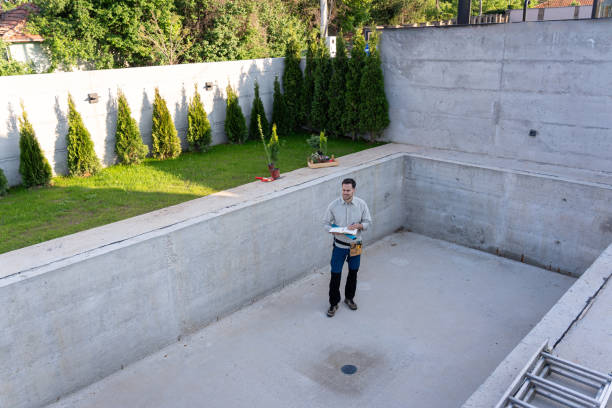 A pool builder from Mooresville Get Swimming Pool Builders by Craft Master Pools and Spas standing inside a completed, empty, concrete pool shell, reviewing plans on a clipboard. The backyard has a lawn and a privacy wall with small trees.