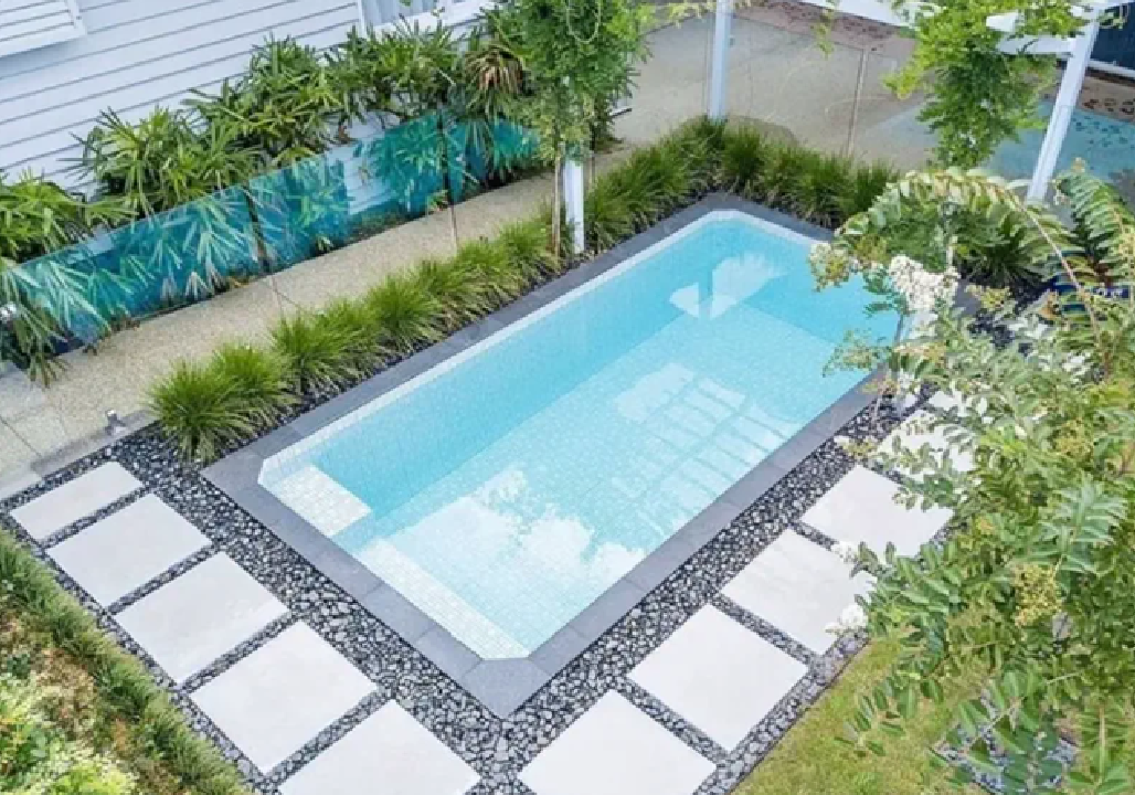 A contemporary rectangular swimming pool is set into a backyard with light-colored stepping stones surrounded by black river stones, and lush green plants and trees. Focus keywords: Mooresville Build A Pool In My Backyard by Craft Master Pools and Spas.