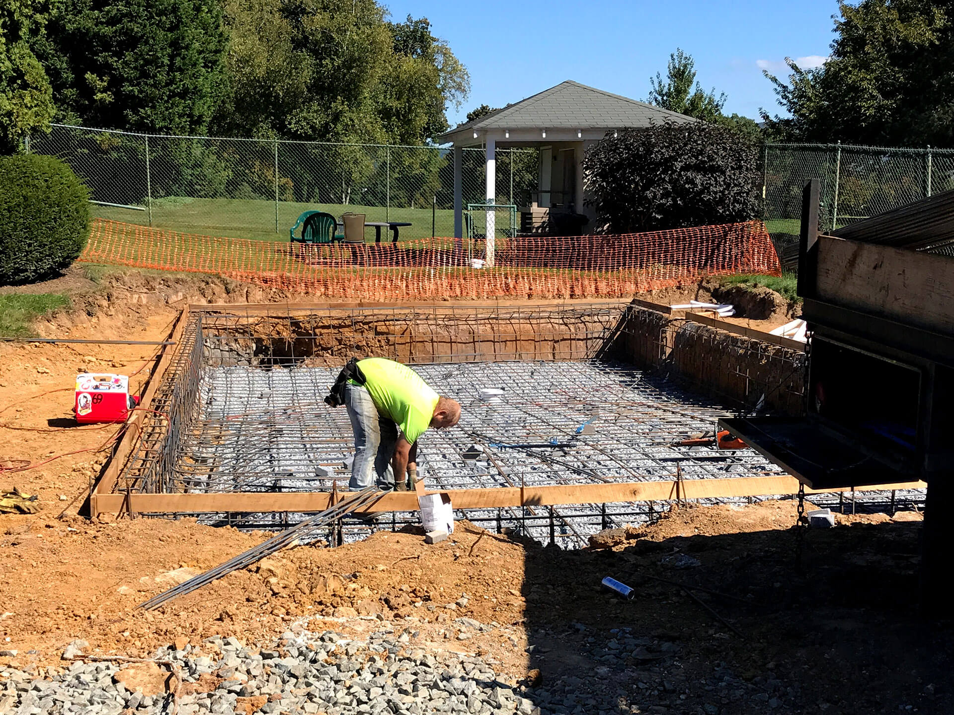Construction in progress by Mooresville Get Swimming Pool Builders by Craft Master Pools and Spas, showing a worker bending over to tie rebar for the pool's structure inside the excavated area, surrounded by wooden forms and soil. A gazebo is visible in the background.