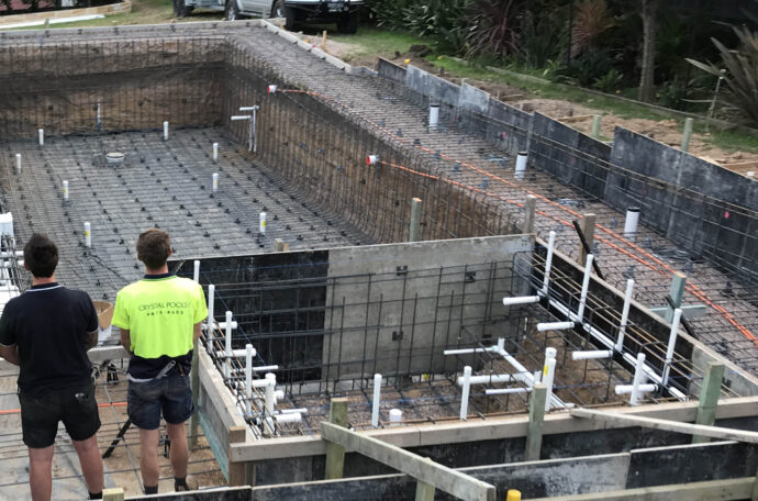 A concrete (gunite) swimming pool under construction, showing the rebar framework and plumbing pipes being installed by Mooresville Swimming Pool Contractors by Craft Master Pools and Spas. Two contractors oversee the complex structure.