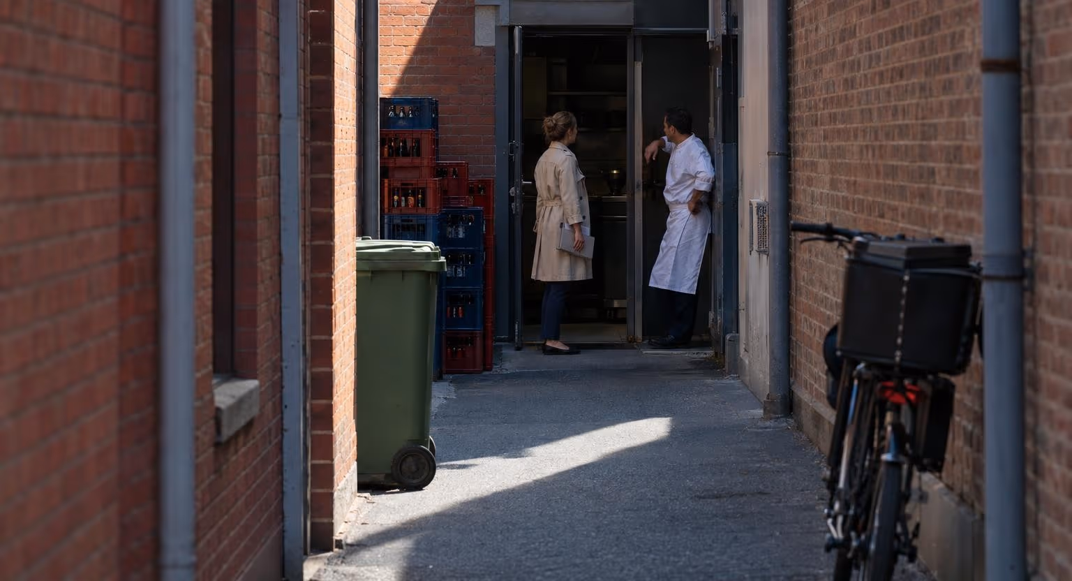 Une femme avec une chemise cartonnée parle à un homme en tenue blanche appuyé à l'encadrement d'une porte, dans une ruelle étroite.