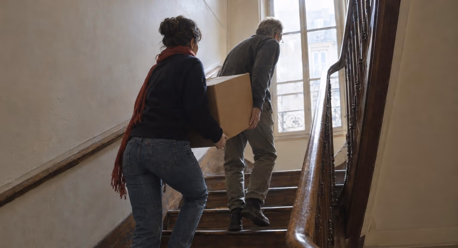 Une jeune femme aide un homme âgé à monter un carton dans l'escalier en bois d'un vieil immeuble, chacun le tenant d'un côté