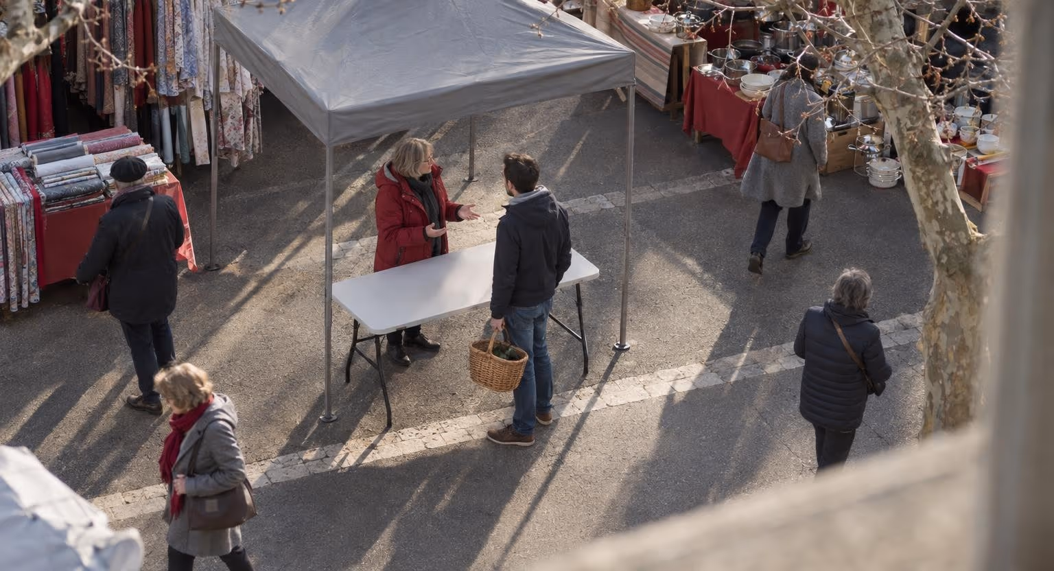 Vu d’en haut, un matin de marché, une femme en parka rouge parle sous un barnum gris avec un homme arrêté devant sa table pliante, un panier à la main.