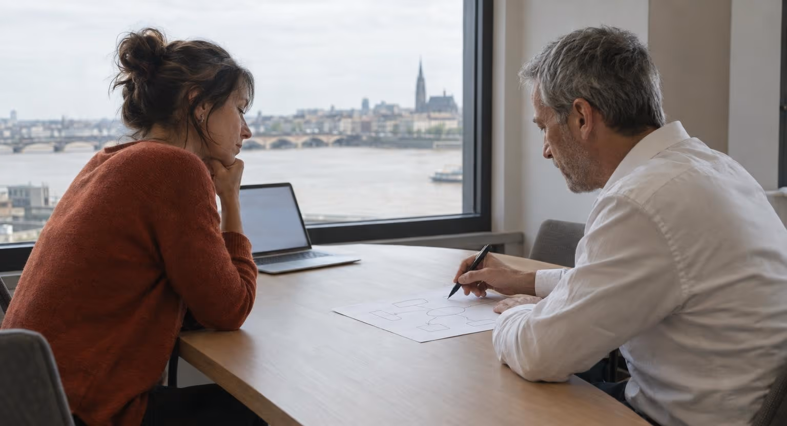 Dans une petite salle donnant sur un fleuve, une femme en pull rouille écoute un homme qui dessine un schéma sur une feuille.