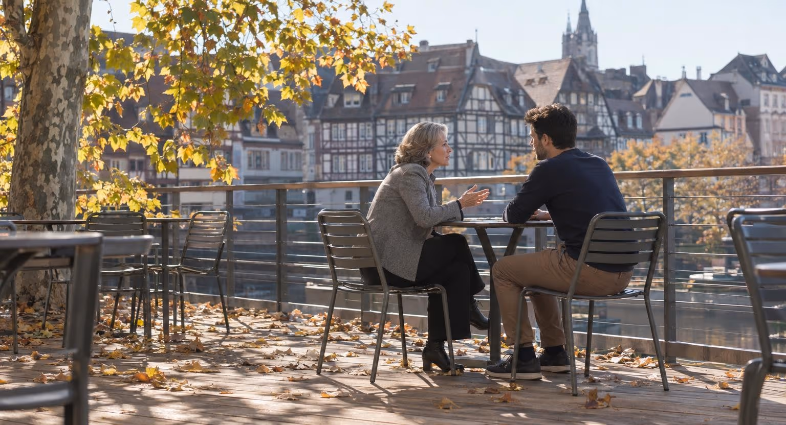 Sur une terrasse en bois, une femme en veste grise parle à un homme plus jeune, attablés à une petite table en métal sous un soleil d’automne.