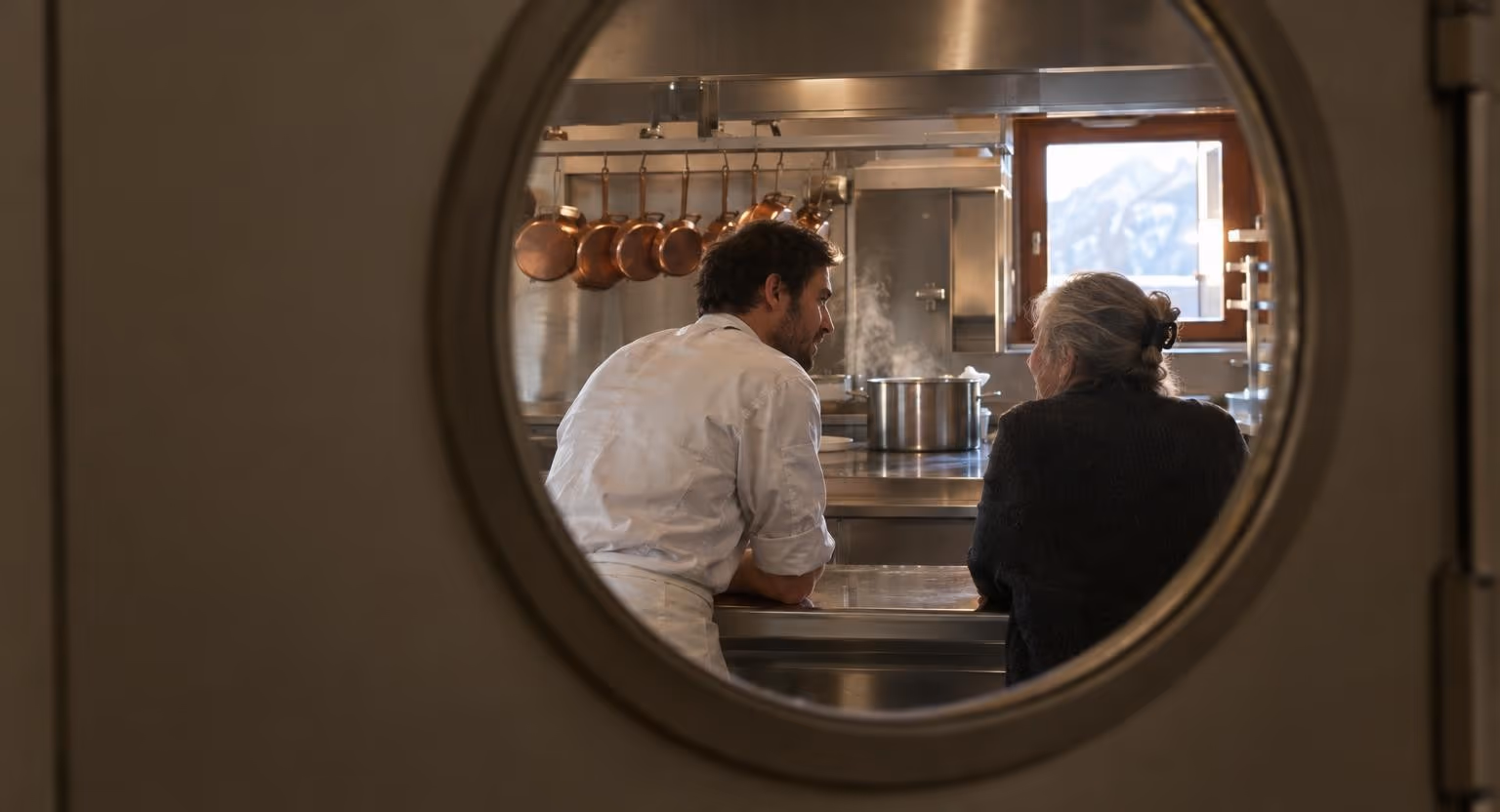 Dans une cuisine de restaurant, un homme en tenue de cuisinier et une femme en gilet sombre discutent, accoudés côte à côte au passe en inox.