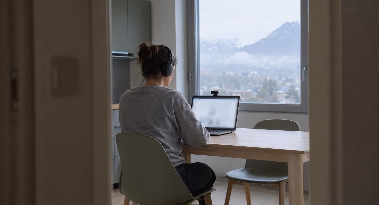 Seule à une table de cuisine vide, une femme en sweat gris, casque sur les oreilles, fait face à un ordinateur portable surmonté d’une petite caméra.
