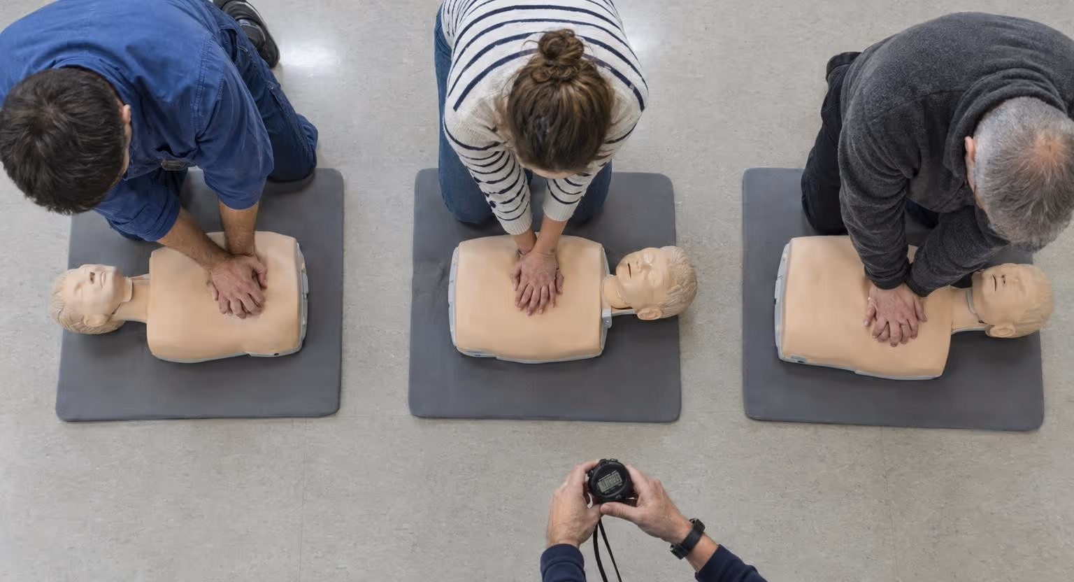 Vues d’en haut, les mains de trois adultes agenouillés pressent la poitrine de mannequins allongés sur des tapis gris.