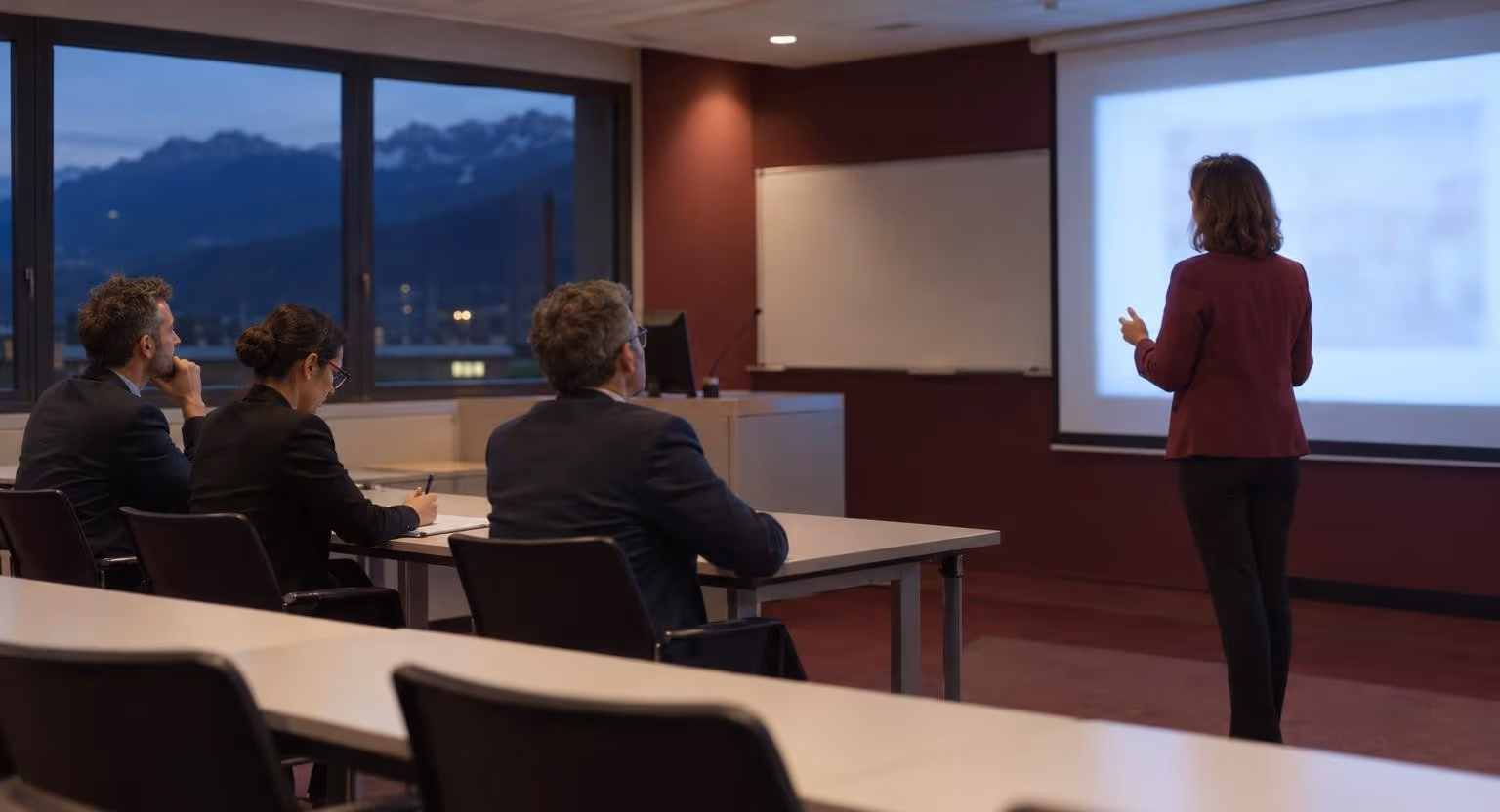 Debout près d’une projection floue, une femme en blazer bordeaux présente un travail à trois personnes assises derrière une rangée de tables.